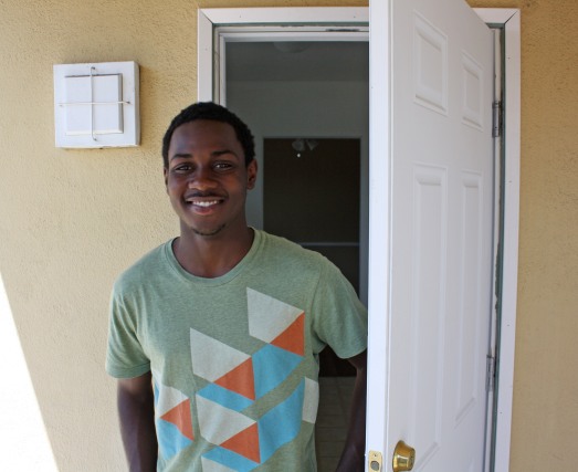 Photo: Sheldon, a young black man, stands smiling in an open doorway. 